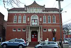 Two-story brick building with arched windows on second floor and small rectangular windows on first, with a red door and pointed facade. A statue stands in front of the right side of the building.