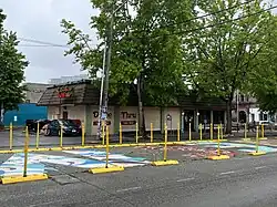 Thin yellow traffic barricades divide the mural from the street, with trees and a Ranchos Bravos tacos store are in the background. The mural is upside-down, with an "M" painted with a white background and splashes of blue, purple, teal, and yellow painted over the M and sometimes into the street. The "S" is filled with thick blobby lettering in red with white outlines over a green background. The "E" has a light blue background with yellow, pink, and purple blobs.