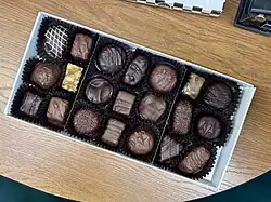 A box of assorted chocolates sits on a wooden table, filled with various shapes and textures including truffles, caramels, and nut clusters in dark and milk chocolate.