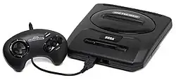 Photograph of an electronic video game console with single controller plugged in the front. The console is encased in black matte plastic that is shaped as a flattened rectangular cuboid with rounded corners. The top has two buttons and a slot for cartridges, while the front has two ports. A black game controller with a black cable is plugged into one of the ports.