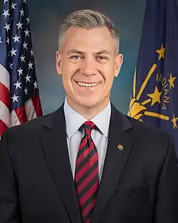Official Senate portrait of Banks smiling in front of the U.S. and Indiana flags, wearing a black suit with gold lapel pin, light blue shirt, and striped black and red tie.