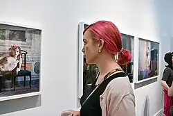 A pink-haired woman closely observes a framed photograph on a white gallery wall.