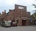 A brick building with an angular façade consisting of stepped brick walls above an entrance porch with a wooden door in a recess. A crucifix and the words "SHOREHAM FREE CHURCH" are on the façade.