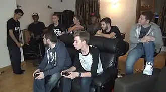 Nepenthez and Calfreezy hold video game controllers on a sofa with the Sidemen sitting behind them
