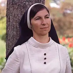 A nun in a white habit with a long dark veil stands outdoors by a tree, gazing calmly into the distance