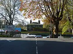 A photograph of the T junction of a road taken from the stalk of the T. A river runs across the top of the T and a small brick cottage can be seen on the far bank.