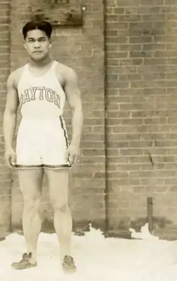An Asian man wearing slippers in a sleeveless shirt and running shorts standing in snow against a brick wall