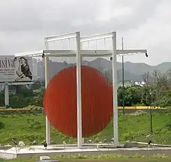 Daytime photo of sky, mountains, vegetation, a billboard, and, in the center of the image, poles with an orange circle in the center