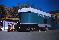 A line of people waiting to enter a green trailer with "movie theater" (kinoteatr) written on it in Russian.