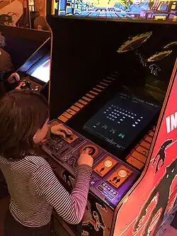 Close up of a young girl playing an arcade video game. The game is in a vertical wooden structure with a visual display unit embedded in the front side. The sides have red panels with artwork depicting jagged silhouettes of monsters over the background of an alien planet surface. A control panel rests below the monitor with a black joystick on the left and a red buttons in the center.