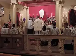 An altar surrounded by altar servers with lit candles, flowers, and a cross in the center, and a rose-colored wall in the back