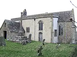 A small stone church seen from the south-east with a porch to the left, a protruding rendered south aisle in the middle, and a short chancel to the right. On the west gable is a bellcote, and in front of the church is a churchyard cross
