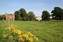 A grassy field, its uneaven surface barely discernable, is bright green under a blue summer's sky. In the foreground are the bright yellow flowers of Rattle. A small brick chapel stands in the rear left corner of the field, and beyond that are a row of mature oaks. A gap in the row of trees gives a view of the frontage of Stainfield Hall