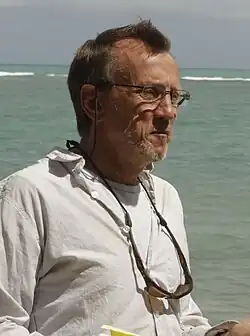 An older man wearing glasses and a light button-up shirt stands near the ocean, with sunglasses hanging from his neck.
