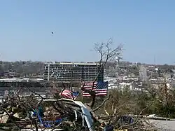 A photo of several downed trees and a waving American flag in the foreground, and the Hilton Convention Center Hotel in the background with shattered windows and other damage