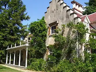 The stepped-gabled west facade of the house,[23] which faces the Hudson River, with the porch – which Irving called his "piazza" – beyond it. It is uncertain why Irving put "1656" at the top of the wall, since the original cottage dates from the 1690s.
