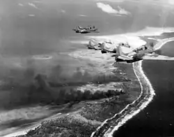 Four propellered aircraft flying over an island. Behind them are two large plumes of black smoke, emanating from the island.