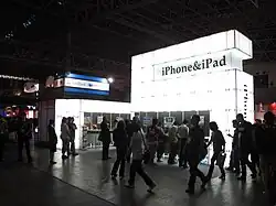 Photograph of a group of people crowded around a large trade show kiosk with a sign that reads "iPhone & iPad". The kiosk is a large structure made of bright white light panels. Demonstration sections surround the lower half of the structure.