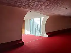 An opening within the curved wall of a flight tube, which leads to a glass-walled passageway that connects with the hotel rooms