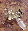A T-bore glass stopcock in a three-way assembly. Two of the outlets end in plain hose adapters while the third ends in a male 14/20 ground glass joint. This stopcock is attached with an easily removed metal spring.