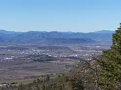 Vegetation in the foreground, with a flat area scattered with buildings farther away. The mesa is far away, with mountains by the horizon.