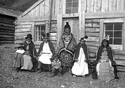 A black-and-white photo of six indigenous people in front of a log cabin
