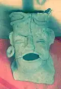A cracked ceramic head with open mouth and closed eyes. The left ear sports an earplug. The forehead is heightened and flattened at the top.