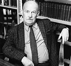 A moustached bald man reclining in a chair in front of a bookshelf.
