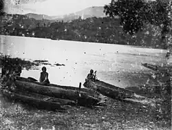 Damaged black and white photo showing two people and log canoes on a lakeshore