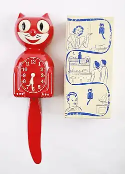 Red cat-shaped wall clock with wagging tail pendulum, displayed next to vintage packaging box