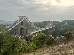 Suspension bridge between two brick built towers, over a wooded gorge, showing mud and water at the bottom. In the distance are hills.