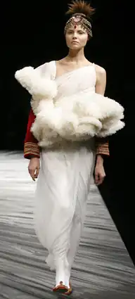 Woman wearing ankle-length slimline ivory gown with large ruffles on the chest and red sleeves, with white tights and red flat shoes