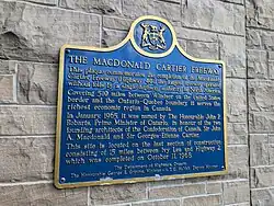 A blue plaque on a stone wall. The plaque has a yellow border, and is mostly rectangular in shape, with the long end oriented horizontally. However, the top side has a camel hump in the centre, with a circle centred at the top of the hump. Inside the circle is an Ontario coat-of-arms. The plaque reads: THE MACDONALD CARTIER FREEWAY This plaque commemorates the completion of the Macdonald-Cartier Freeway (Highway 401), the longest freeway operated without tolls by a single highway authority in North America. Covering 820 kilometres (510 mi) between Windsor on the Canada–US border and the Ontario–Quebec boundary, it serves the richest economic region in Canada. In January 1965, it was named by The Honourable John Robarts, Premier of Ontario, in honour of the two founding architects of the Confederation of Canada, Sir John A. Macdonald and Sir George-Étienne Cartier. This site is located on the last section of construction, consisting of 15 miles between Ivy Lea and Highway 2, which was completed on October 11, 1968.