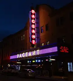 Photographed from an oblique angle, the Pageant's façade is lit by two perpendicular signs (in purple, yellow, and red) displaying its name, as well as a small marquee advertisement