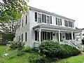 A white colonial frame house with a wide single story front porch. The main rectangle of the house has extensions to the rear.