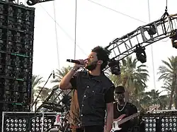 The Weeknd wearing a button-up, sleeveless shirt covering a normal black t-shirt. He is performing on a stage, singing into a microphone, with palm trees in the background.