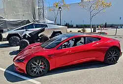 A stainless steel clad ATV between a Tesla Cybertruck and a second generation Tesla Roadster in a parking lot during a Tesla event in 2020.