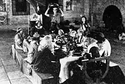 Black and white film still showing actors in medieval costumes seated at a banquet table in a grand hall, while attendants serve food and drink.