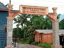 A concrete arch opens into a road bordered by lush vegetation, bearing a sign reading "WELCOME TO OTTA VILLAGE ORAUKWU / BUILT BY IFEADIGO AGE GRADE".