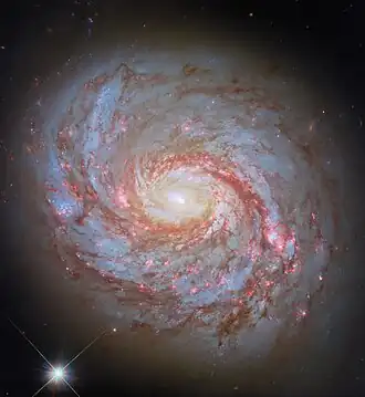 "A close-up of a spiral galaxy, seen face-on. Its center is a bright glowing From the sides of the galaxy's core emerge spiral arms which wind through the round disc of the galaxy, filled with shining pink spots where stars are forming and more dark-red dust. Some faint stars can be seen around the galaxy, as well as a particularly bright star in the lower left of the image."