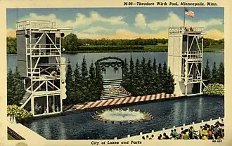 Color postcard depicting a swimming pool surrounded by a seating area, and a stage with diving platforms on both ends