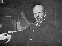 A black and white photo of a bald man with a short beard wearing a dark coat