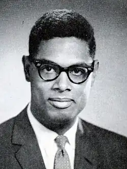 A dark-haired man, wearing glasses and a suit and tie, looks into the camera