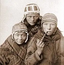 A sepia-toned photograph of three Sámi women wearing traditional caps and clothing, with one woman holding a pipe, taken around the 1890s.