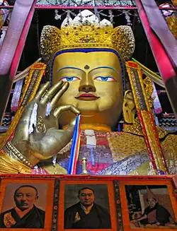 Seated Maitreya Buddha statue inside Tashilhunpo Monastery, showing a gold-colored face, ornate crown, raised right hand, and richly decorated surroundings, with framed portraits beneath the statue.