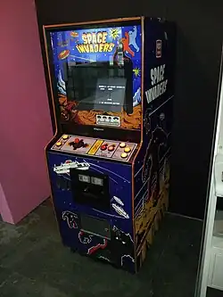 A vertical wooden structure with a visual display unit embedded in the front side. The sides have dark blue panels with artwork depicting flying saucers and jagged silhouettes of monsters over the background of an alien planet surface. Above the display monitor and on the side panel reads: "Space Invaders". A control panel rests below the monitor with a red joystick on the left and two red buttons on the right side.