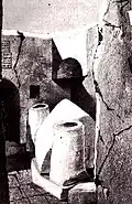 Black-and-white photograph showing an open-air scene of ruins with a white, peaked centotaph in between two short white columns