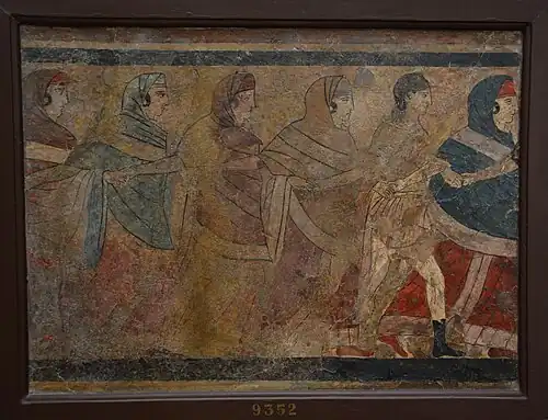 Women dressed in bright blue, red, tan, and black dresses with head coverings dancing in a circle while holding hands. Periodically there is a man in all white with an instrument. They all share they same sorrowful look.