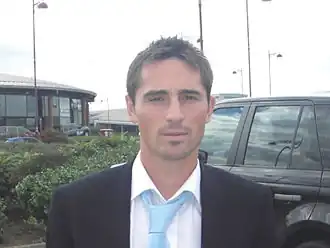 The head and shoulders of a man, wearing a white shirt, blue tie and black blazer