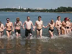 Eight topless women and two barechested men standing in knee-depth water and holding hands, all of whom are shirtless.